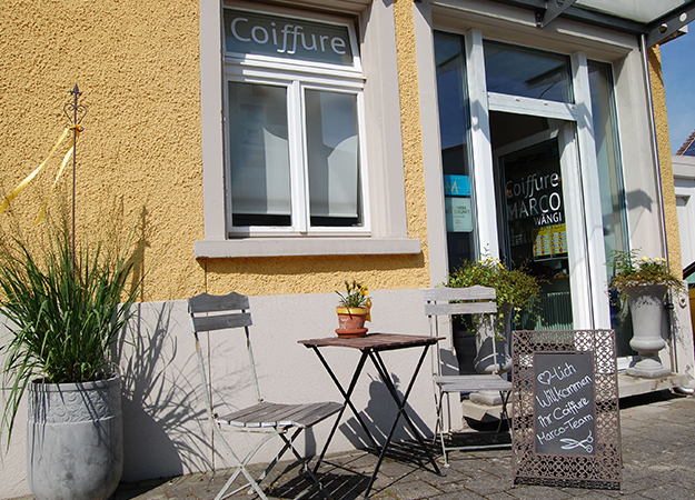 Außenansicht eines gelben Gebäudes mit Eingang zu einem Friseursalon und Sitzgelegenheiten im Freien.