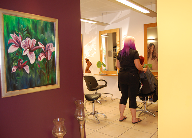 Das Interieur eines Friseursalons: Eine Friseurin mit lila Haaren schneidet einer Kundin vor einem Spiegel die Haare; an der Wand hängt ein Blumengemälde.