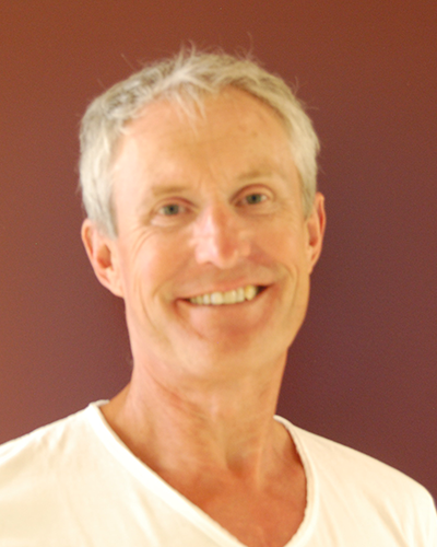 Lächelnder, grauhaariger Mann im weißen Hemd; vor einem rotbraunen Hintergrund.