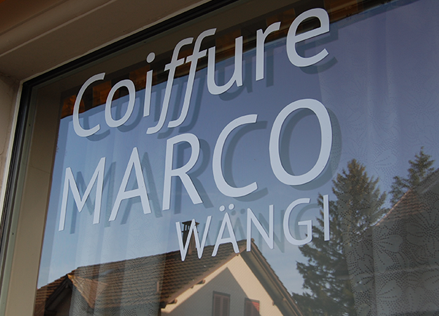 Weißer Schriftzug an einem Fenster: „Coiffure MARCO WÄNGI“. Spiegelungen von Gebäuden und blauem Himmel.