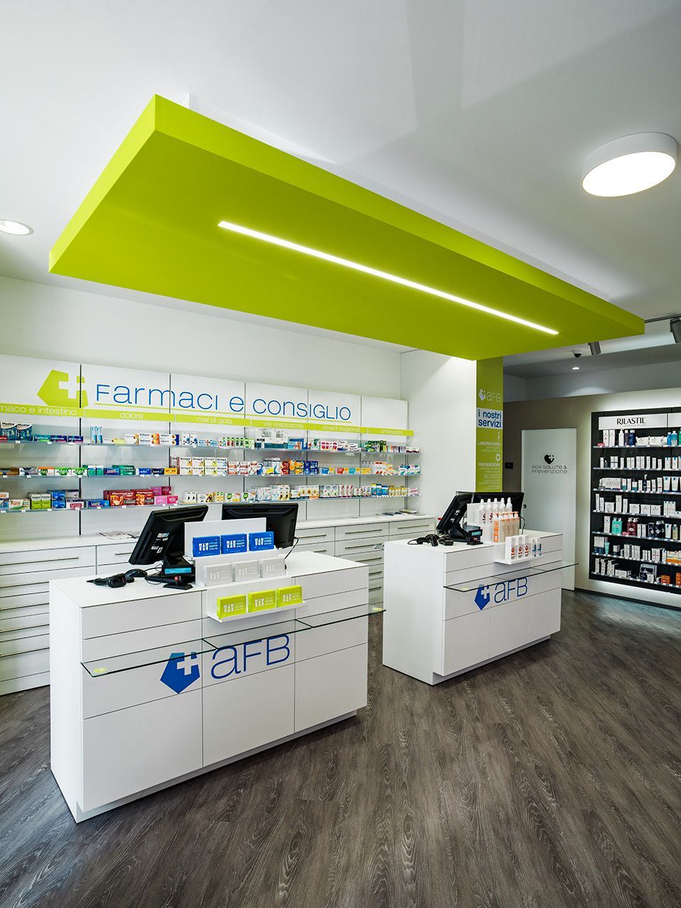 Interno di una farmacia moderna con banconi bianchi, scaffali pieni di prodotti e un soffitto con accenti di verde brillante.