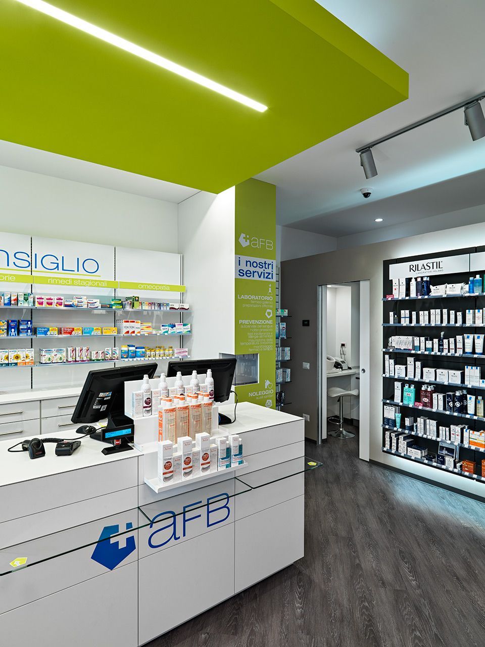 Interno di una farmacia con bancone bianco, scaffali pieni di prodotti e accenti sul soffitto di un verde brillante.