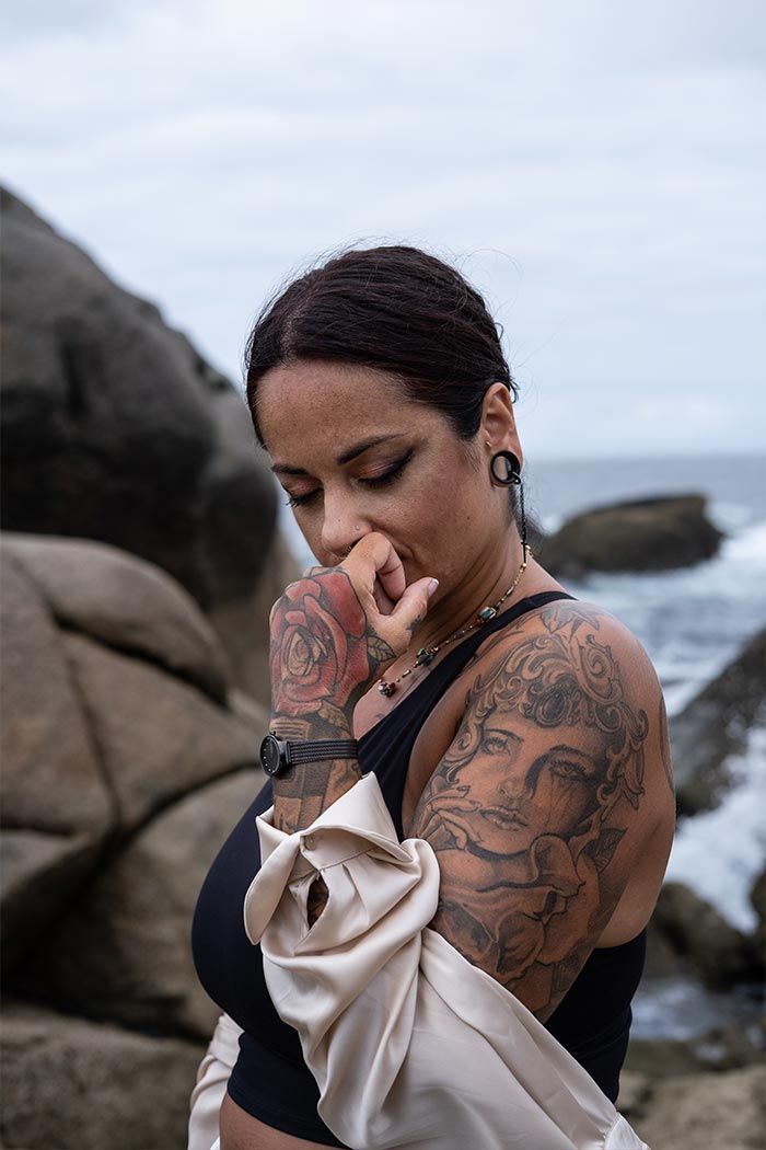 Una mujer con tatuajes en los brazos está parada en una playa rocosa cerca del océano.