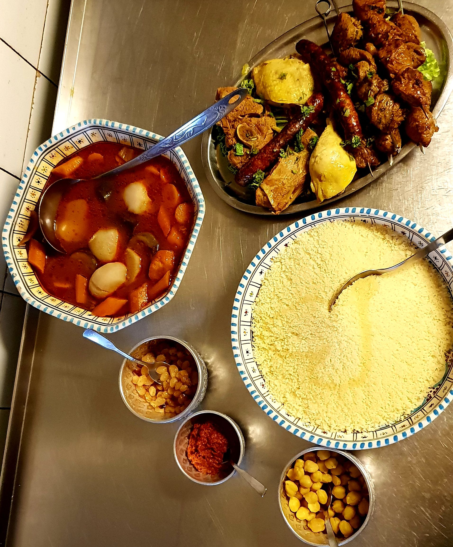 Couscous et ingrédients variés disposés dans des bols bleu et blanc, accompagnés de brochettes de viande et d'épices.