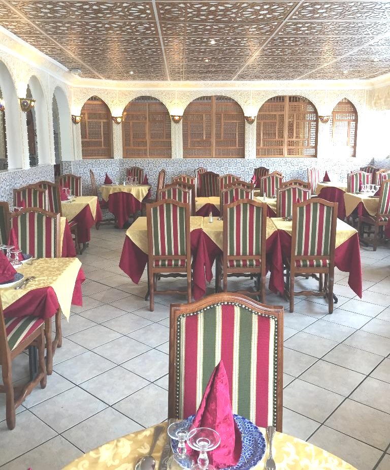 Intérieur d'un restaurant avec tables dressées pour le repas ; plafond orné, fenêtres cintrées, chaises rayées.