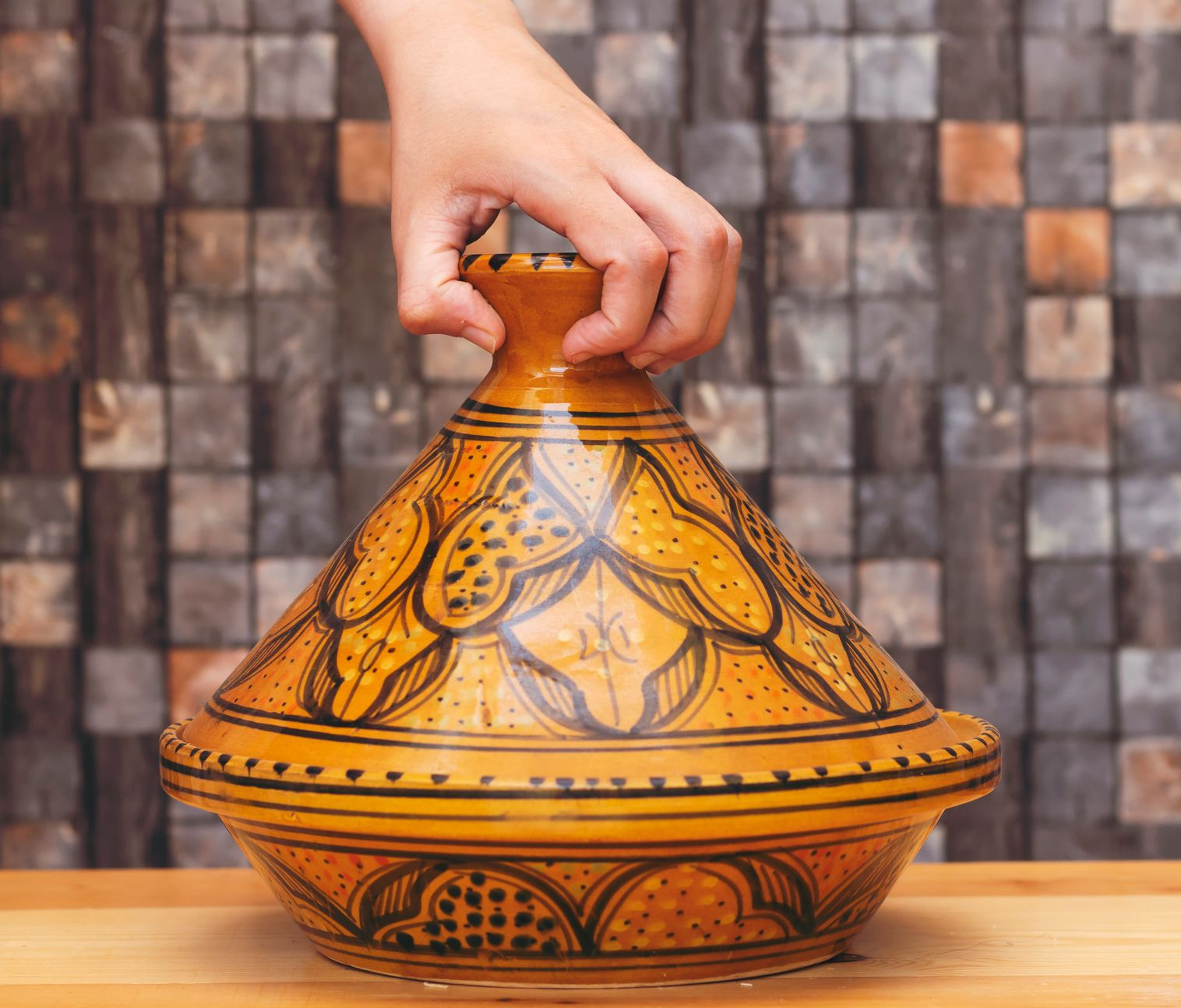 Main soulevant le couvercle d'un tajine décoratif jaune et marron posé sur une surface en bois.