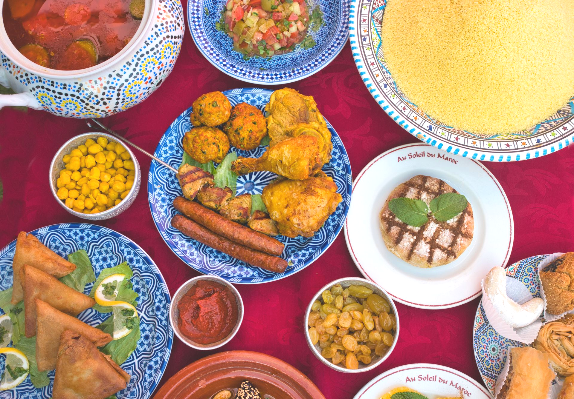 Vue aérienne d'un festin marocain sur une nappe rouge. Au menu : couscous, soupe, viandes et pâtisseries.