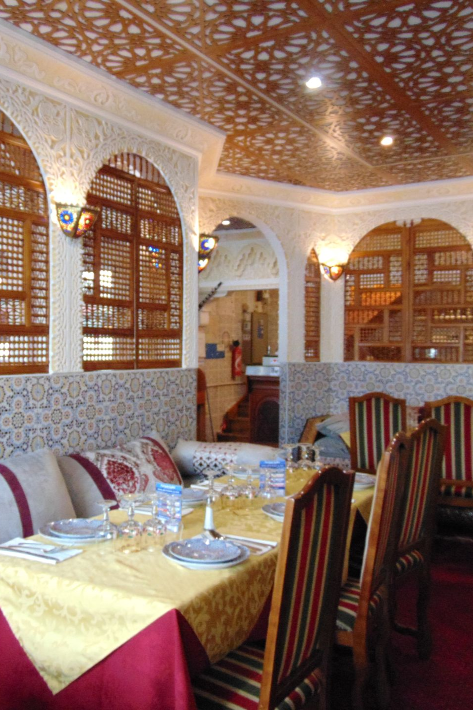 Intérieur de restaurant avec plafond orné et stores en bois. Table dressée avec une nappe jaune, chaises à rayures rouges.