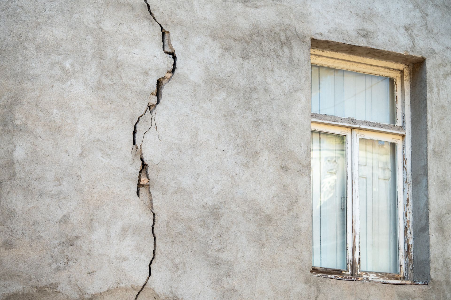 Mur gris avec une large fissure verticale, à côté d'un cadre de fenêtre blanc défraîchi.