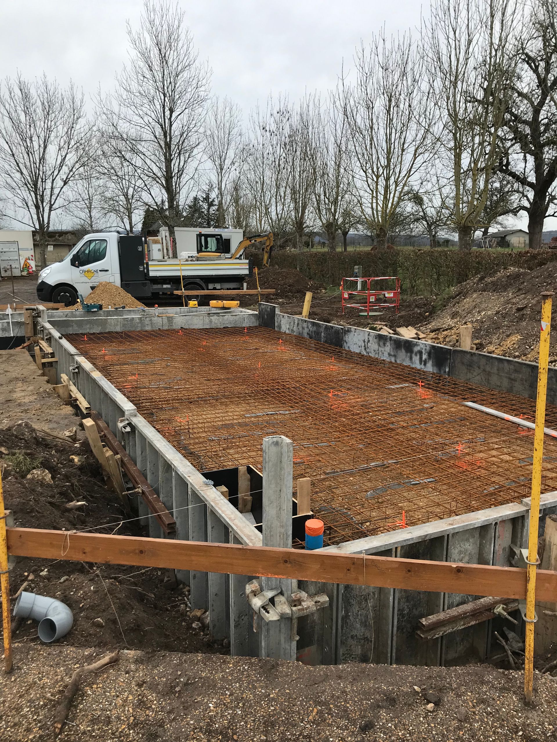 Chantier : coffrage rectangulaire en béton armé, terre et tuyaux. Camion blanc et arbres en arrière-plan.