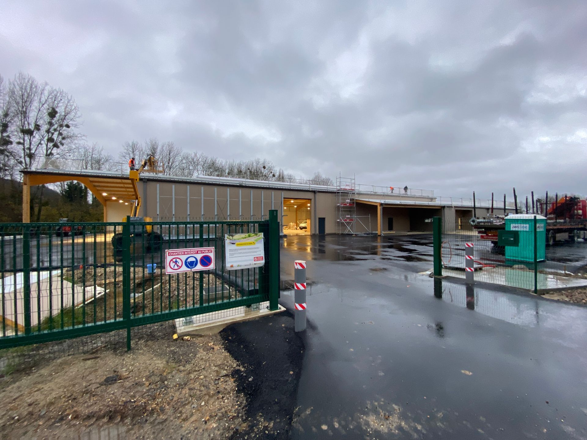 Chantier de construction avec un bâtiment partiellement construit, ciel couvert, portail vert et toilettes portables.