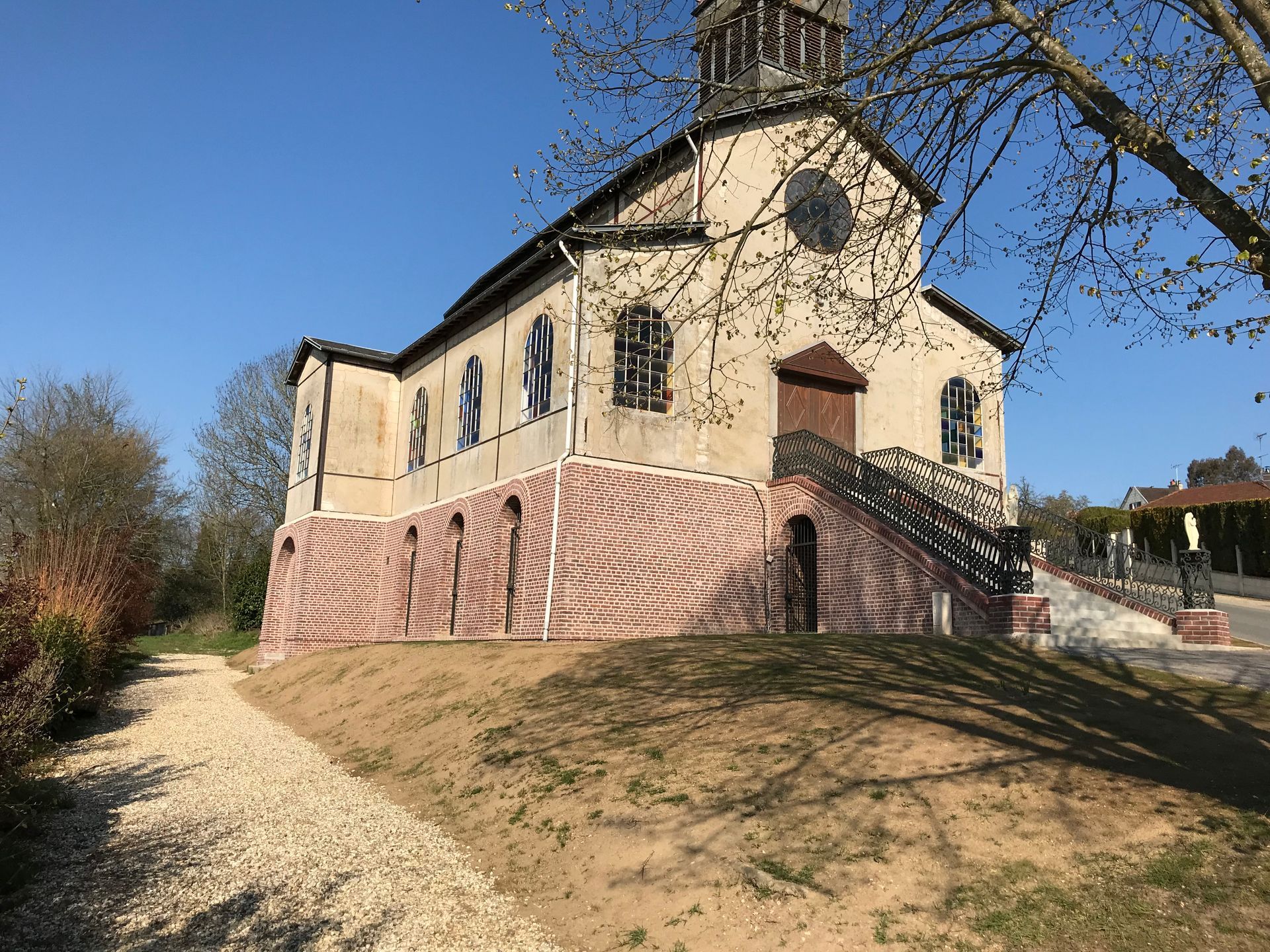 Église sur une légère colline, avec des fondations en pierre, des murs beiges et une porte brune ; chemin au premier plan.