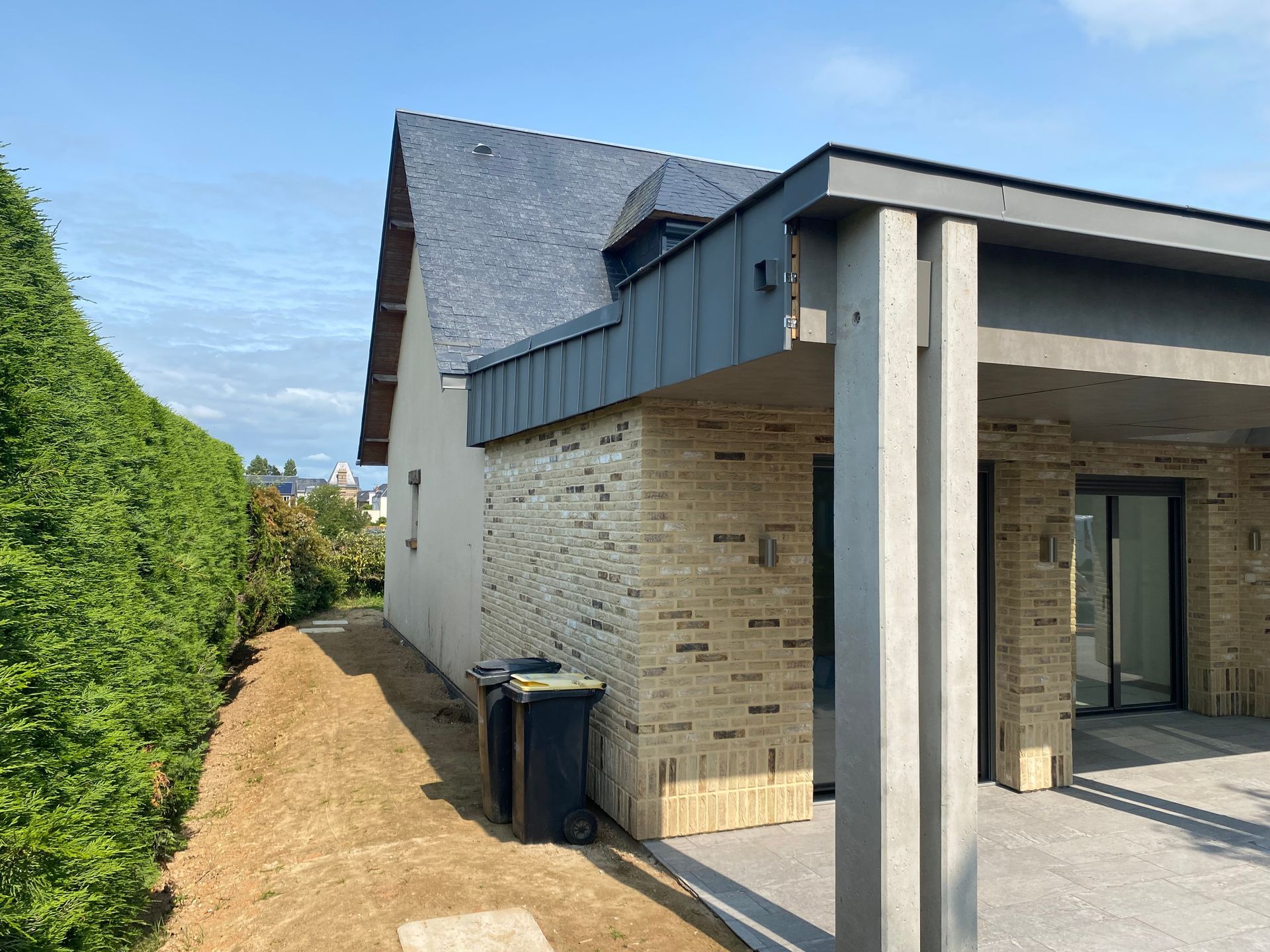 Maison moderne avec façade en briques, abri voiture et haute haie sur le côté, sous un ciel bleu.