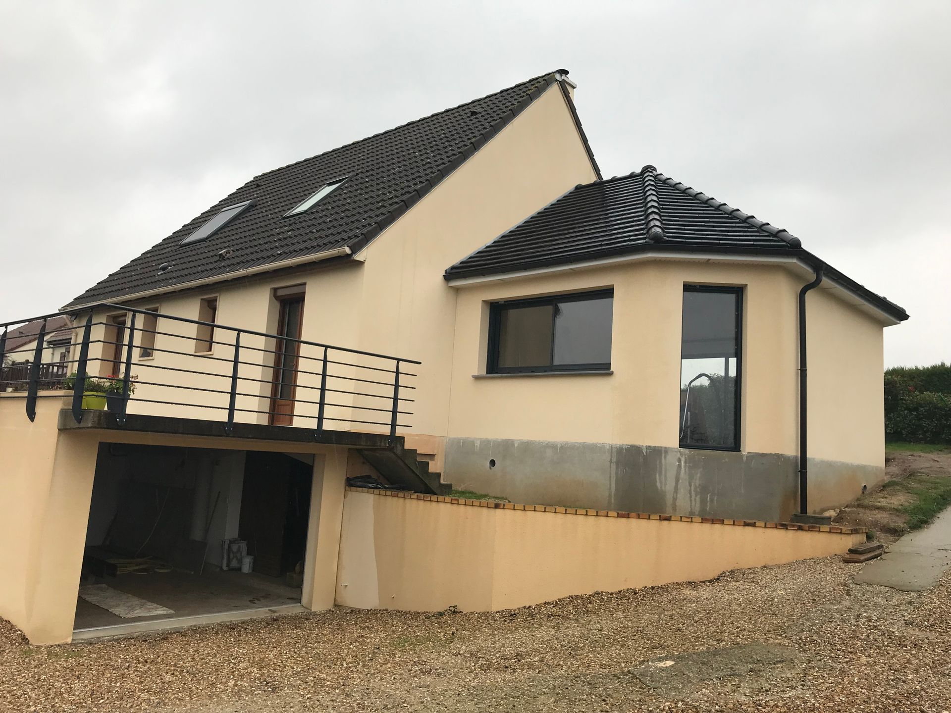 Maison beige au toit noir, garage et escalier menant à un balcon. Ciel nuageux.