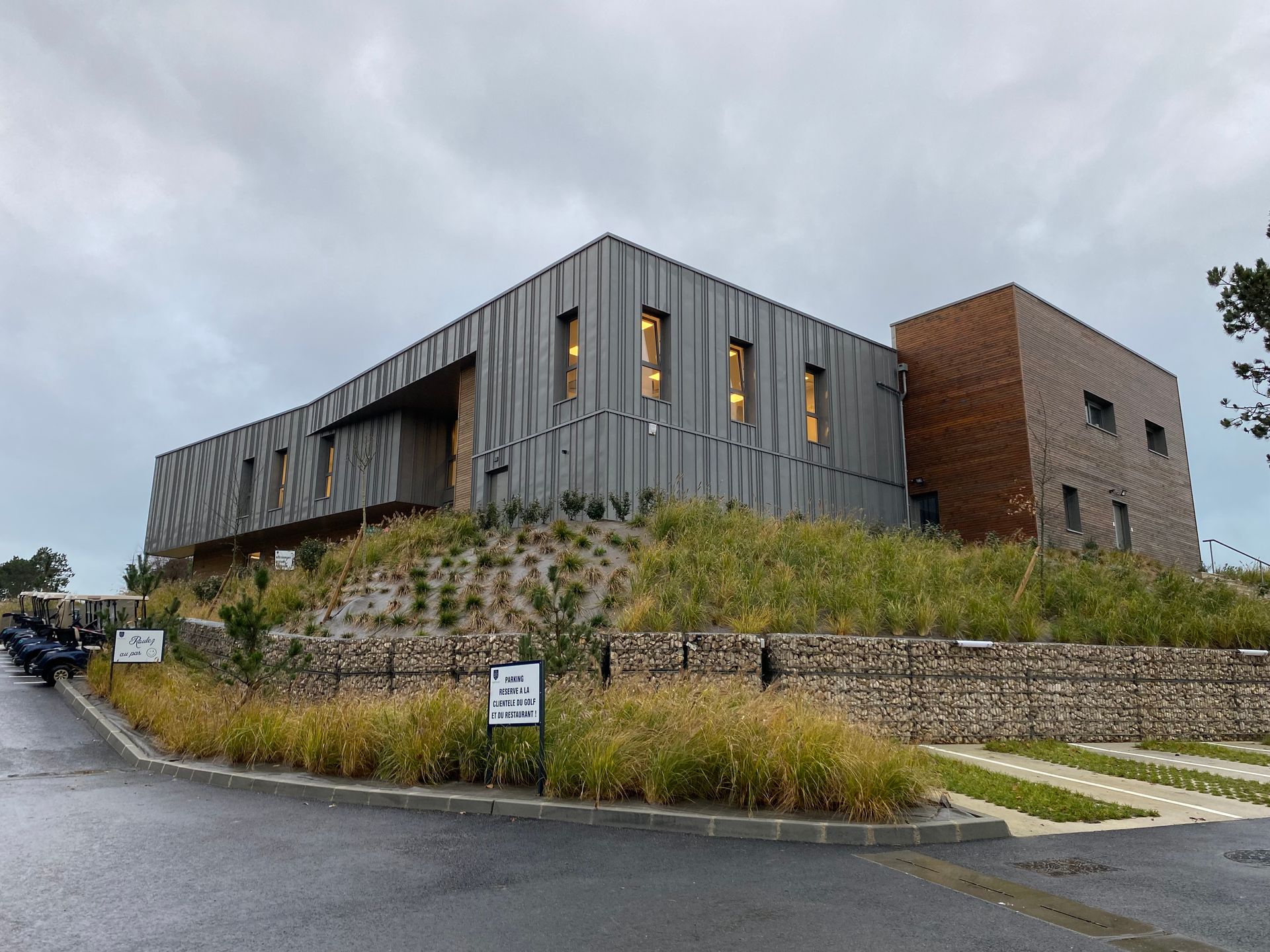 Bâtiment moderne à la façade en métal texturé et en briques, sur une petite colline, sous un ciel couvert.