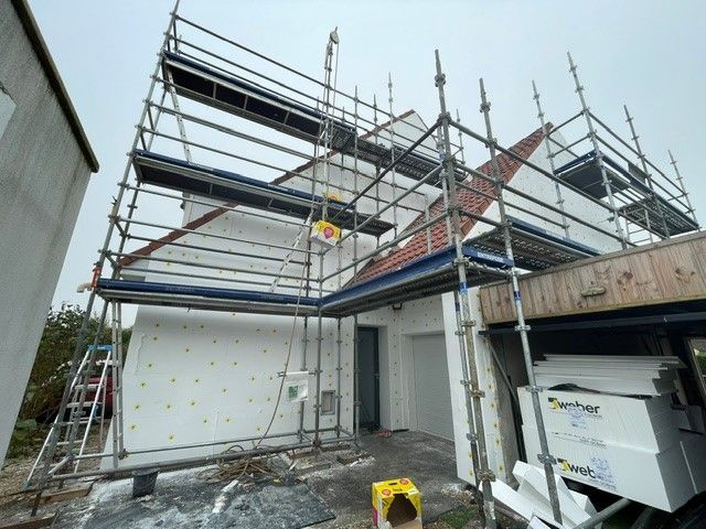 Des échafaudages entourent une maison en construction ; on aperçoit de l'isolant en mousse blanche sur les murs extérieurs.