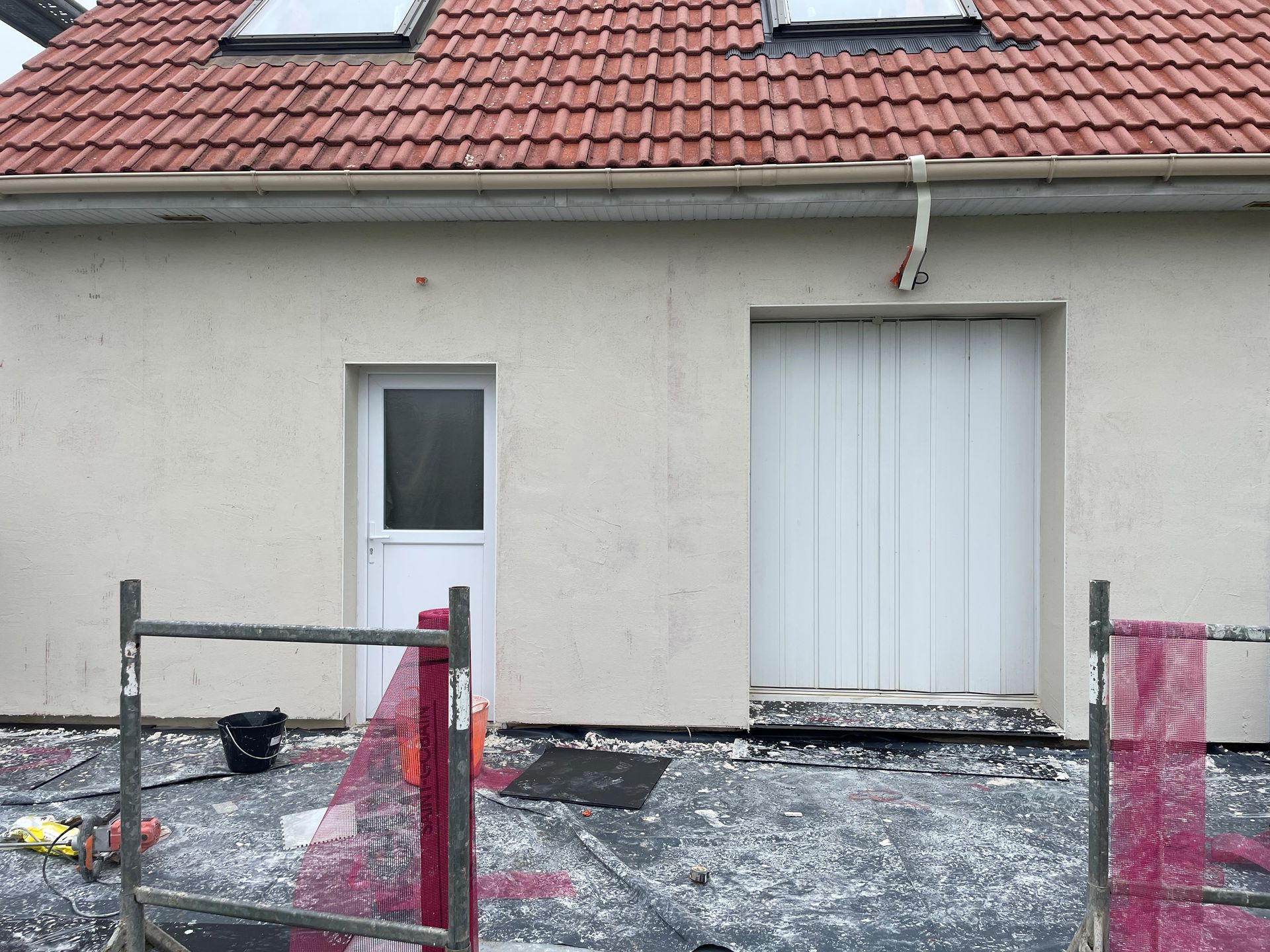 Extérieur d'une maison en construction, avec un toit de tuiles rouges, des murs blancs et une porte blanche.