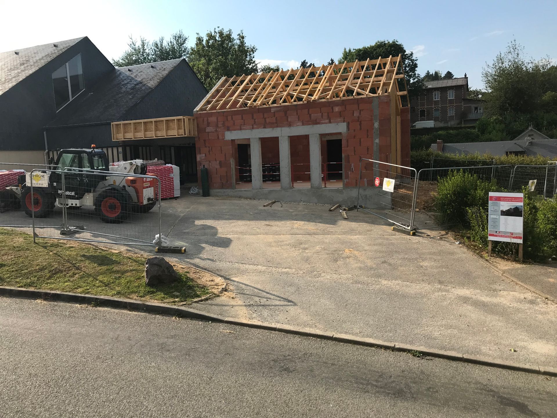 Chantier de construction : bâtiment à charpente apparente, tracteur, matériaux, terrain gravillonné.
