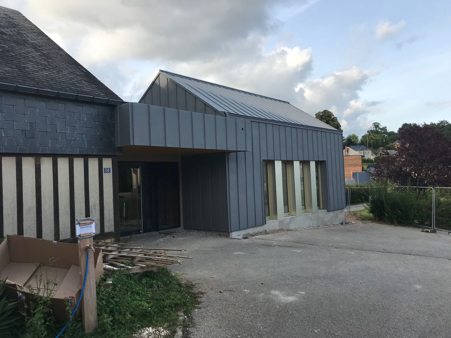 Extérieur du bâtiment en construction avec bardage et toiture en métal gris, béton et fenêtres.