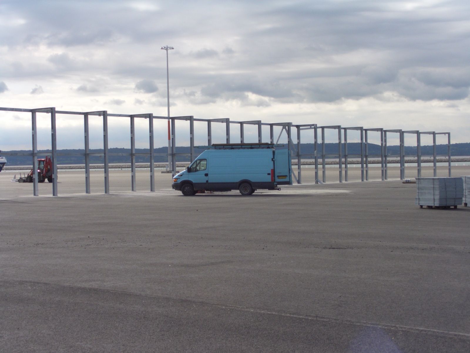 Une camionnette bleue garée parmi des structures métalliques sur un terrain gris et pavé, sous un ciel nuageux.