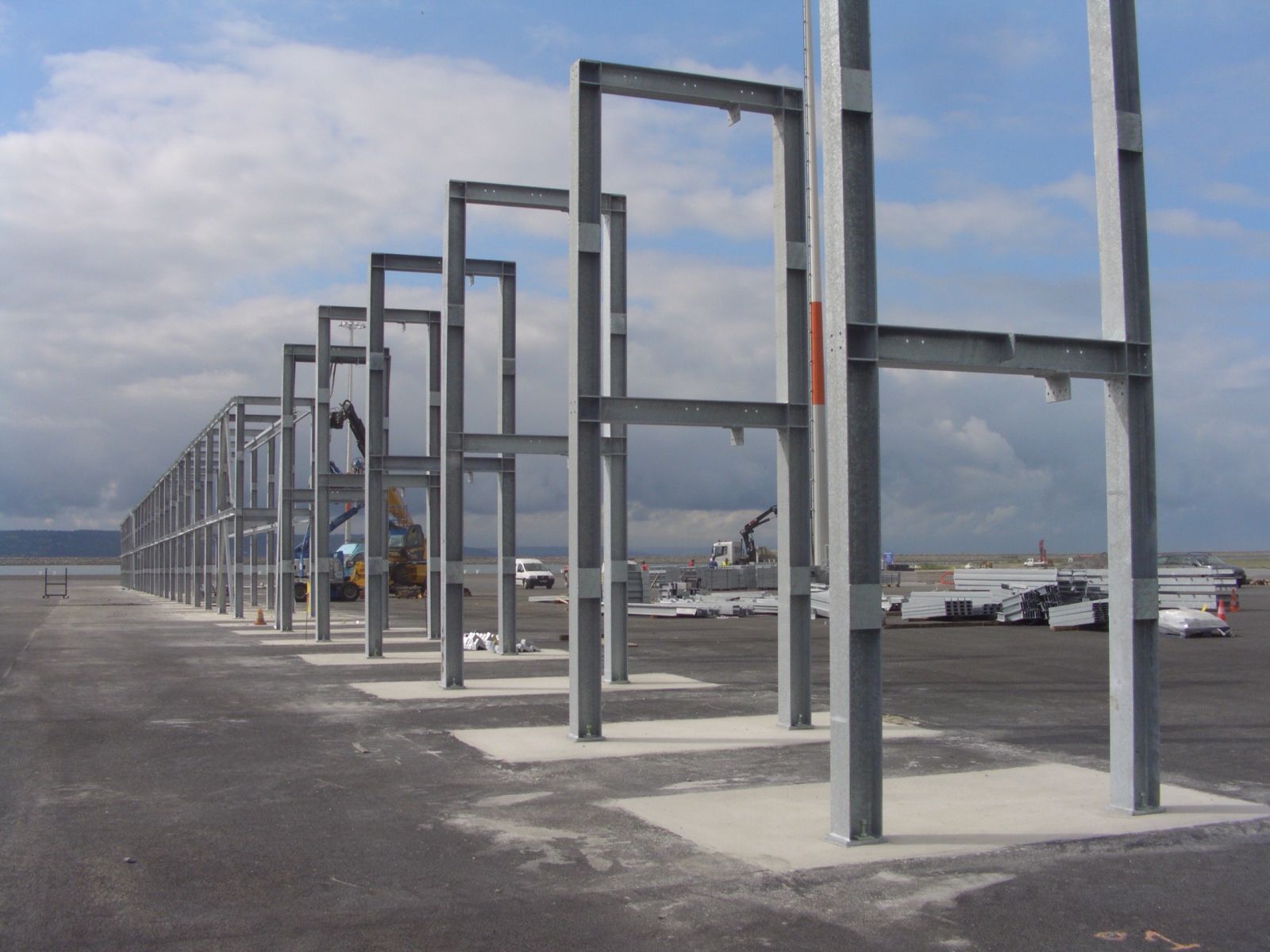 Structure métallique d'un bâtiment en construction, sur une zone pavée grise, avec des équipements au loin en arrière-plan.