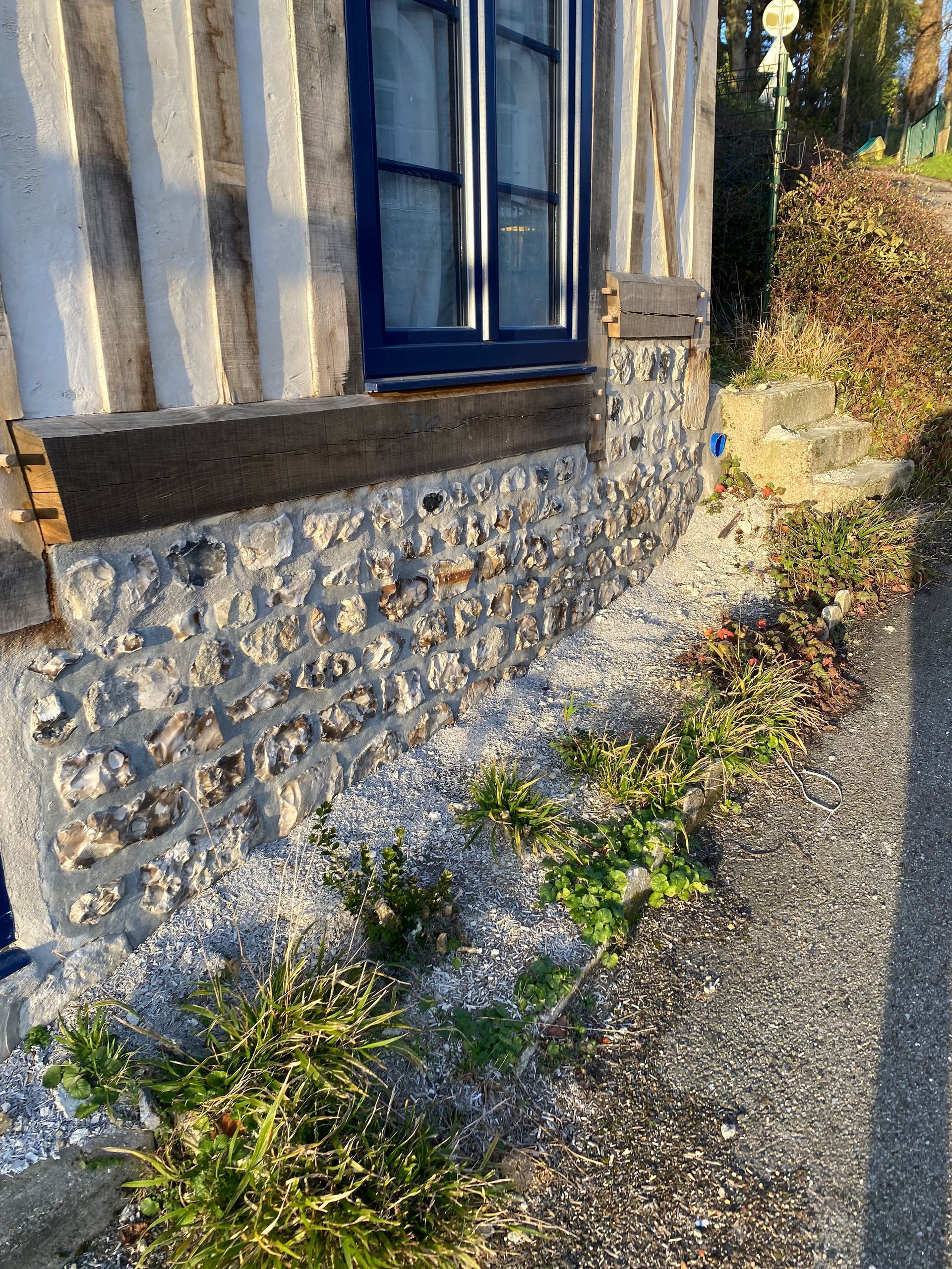 Bâtiment en pierre et en bois, avec une fenêtre, une boîte aux lettres et des plantes en bord de route.