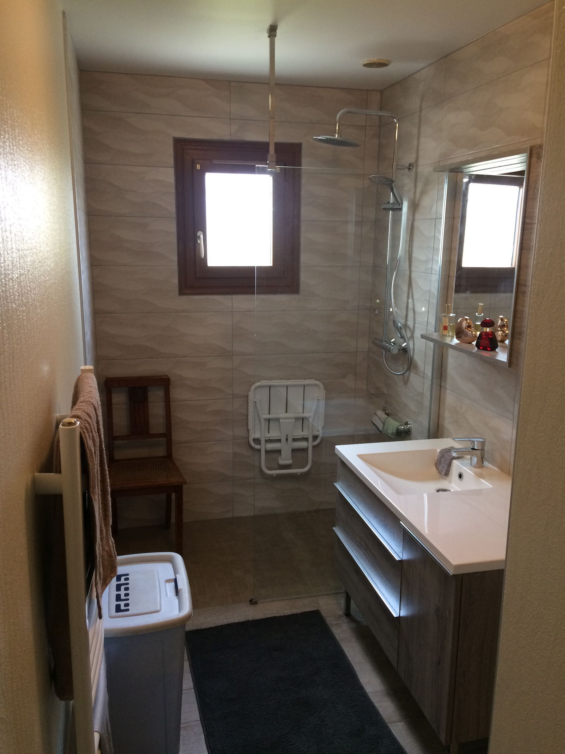 Salle de bains avec douche, lavabo, toilettes et petite chaise. Carrelage beige, tapis foncé et fenêtre.