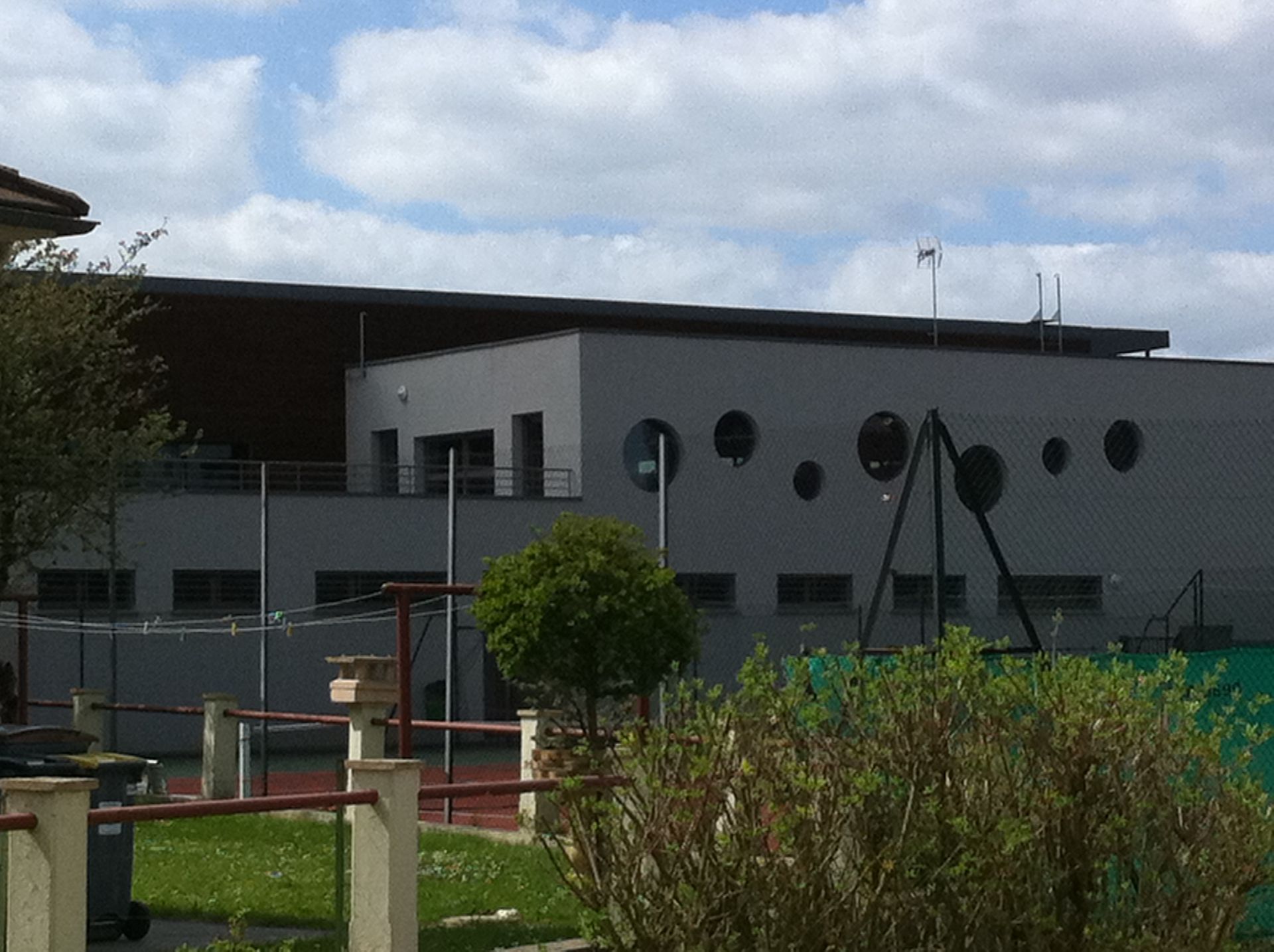 Bâtiment gris avec des fenêtres rondes, une extension de toit marron et des équipements de jeux à l'avant.