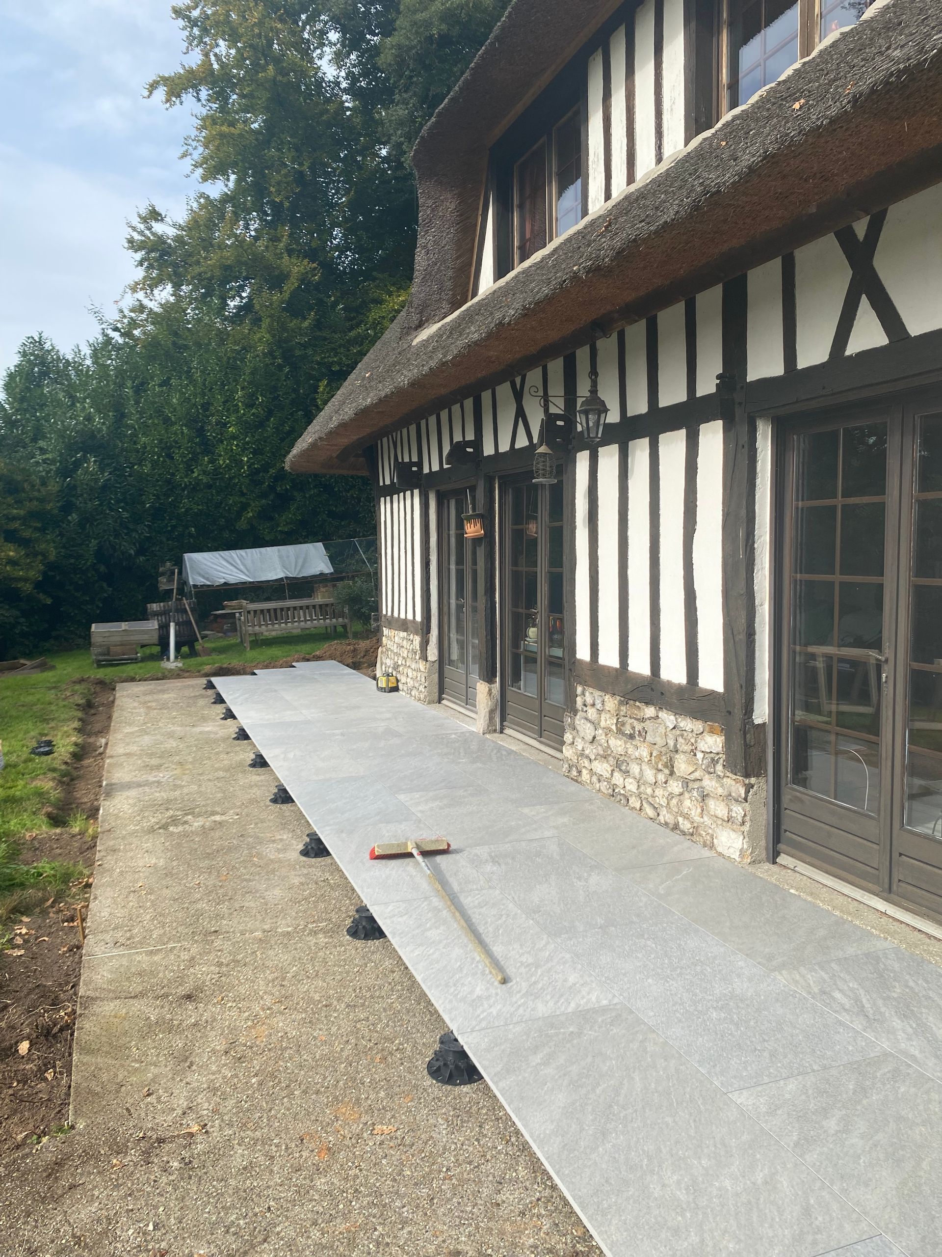 Extérieur d'une chaumière avec une terrasse grise en construction. Un râteau est posé sur la terrasse.