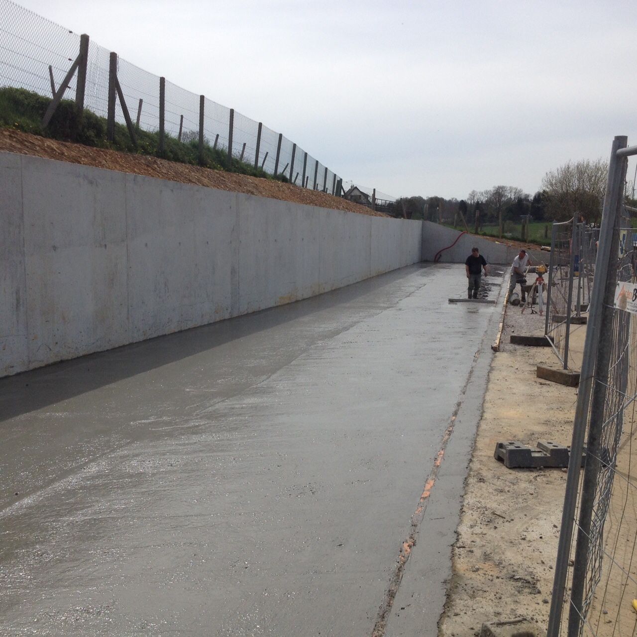 Chantier de construction où du béton frais est lissé ; un haut mur de béton et une clôture délimitent la zone.