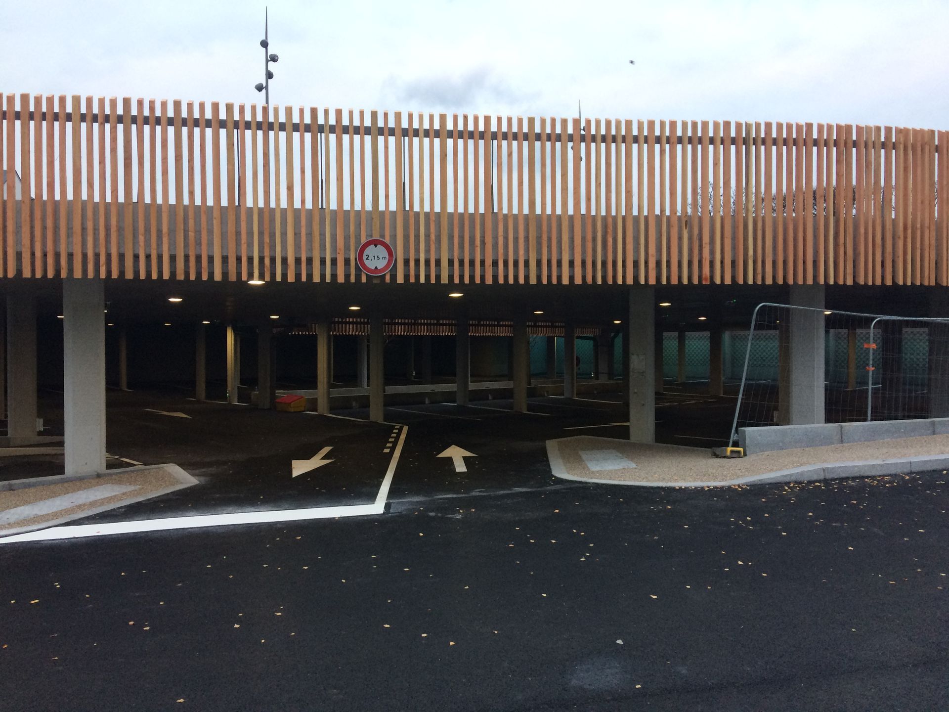 Aire de stationnement sous une structure en lattes de bois, avec voie d'entrée marquée par des flèches.