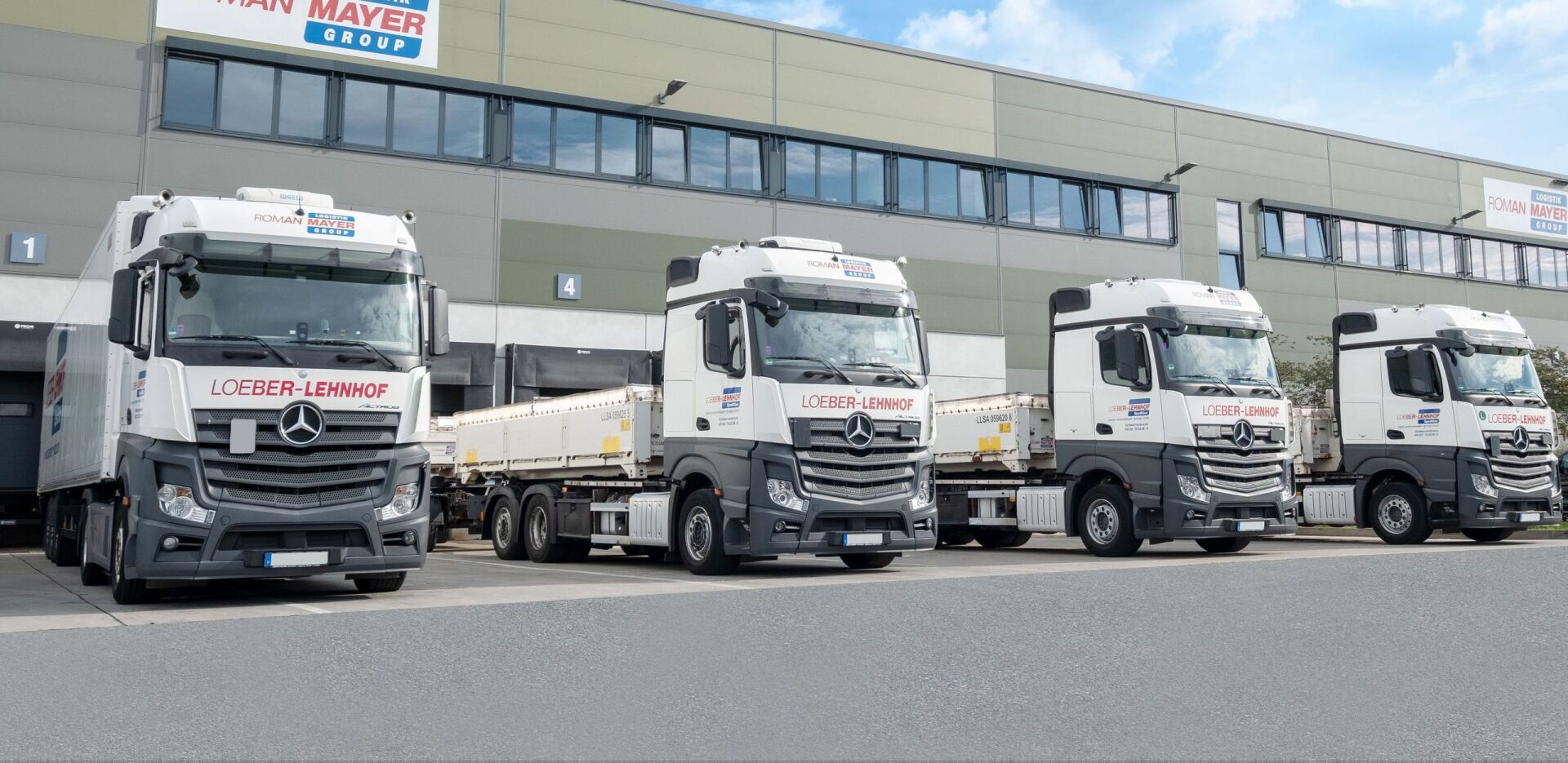 Eine Reihe von vier weißen Mercedes-Benz Sattelzügen parkt vor einem modernen Industriegebäude.