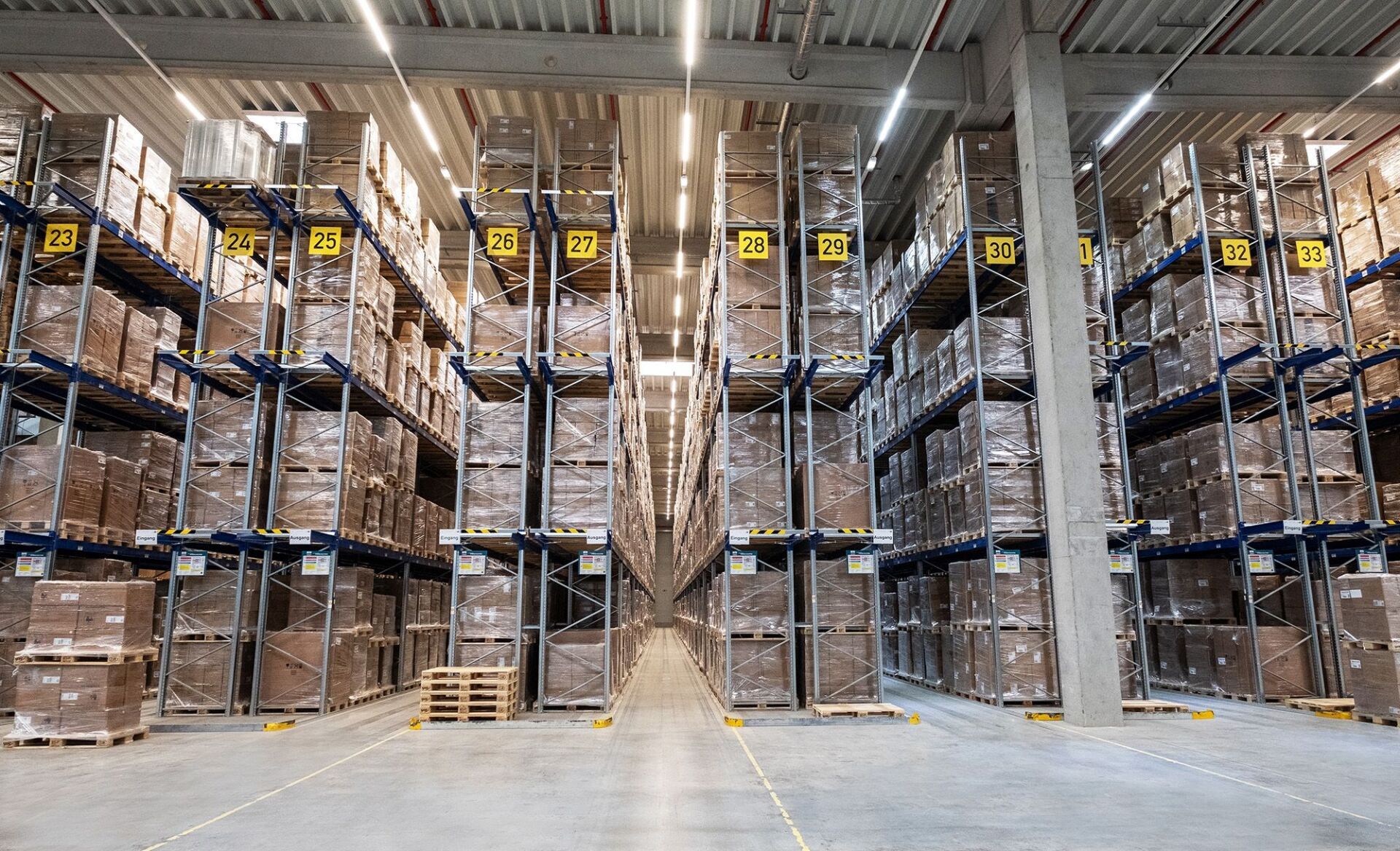 Hohe Industrieregale in einem Lagerhaus, gefüllt mit gestapelten Kisten, getrennt durch einen breiten Betongang.