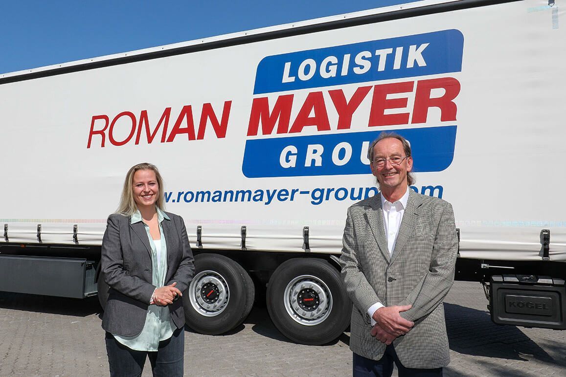 Eine Frau und ein Mann stehen lächelnd vor einem weißen Logistik-LKW-Anhänger der Roman Mayer Group.
