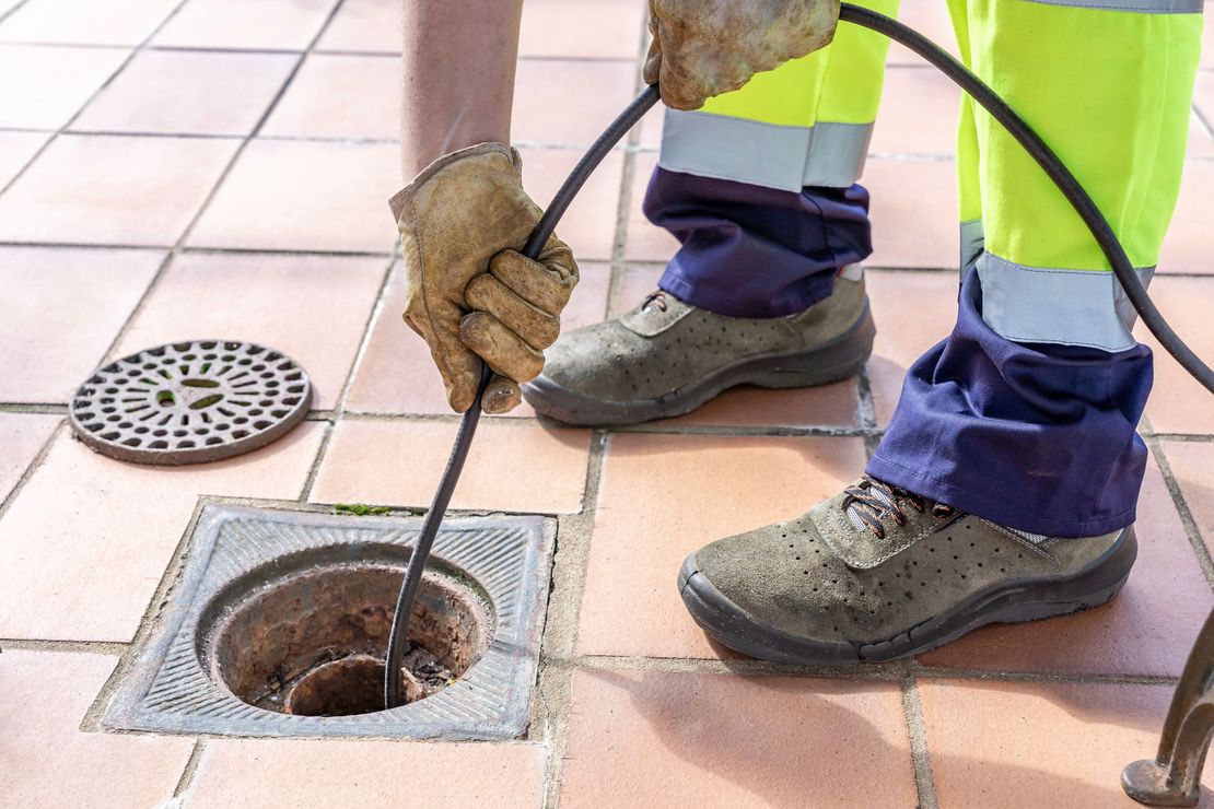 Un ouvrier utilise une longue tige pour déboucher une canalisation sous un carrelage, ses bottes et ses gants étant visibles.