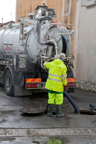 Un ouvrier en ciré jaune fluo manipule un tuyau d'un camion aspirateur à côté d'un bâtiment.