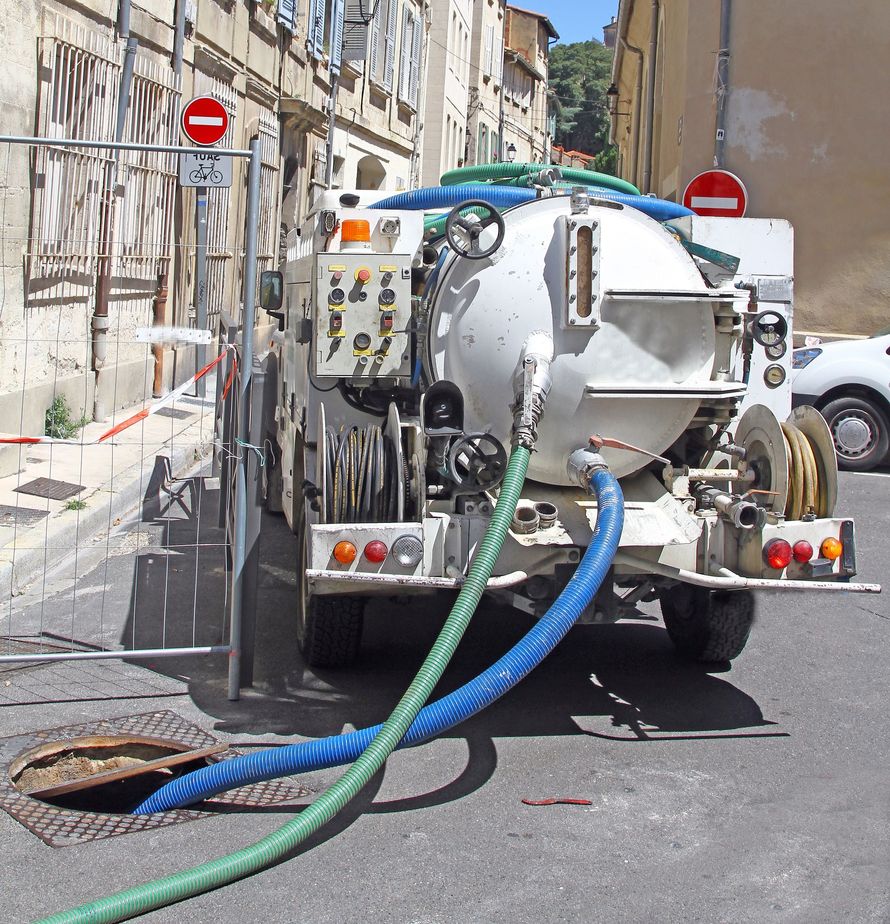 Camion-citerne blanc avec tuyaux nettoyant une rue étroite entre de vieux bâtiments.
