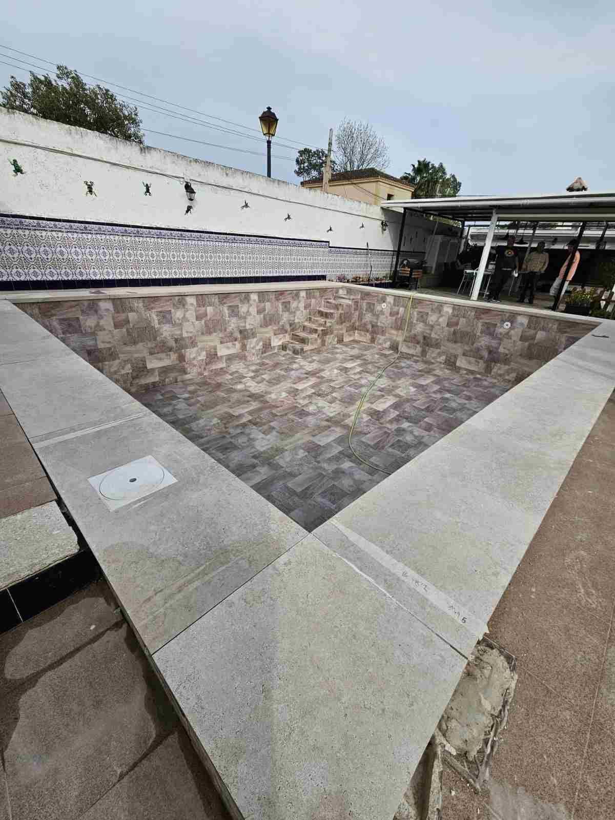 Fuente vacía de azulejos en un patio exterior, rodeada de muros blancos y con un edificio al fondo
