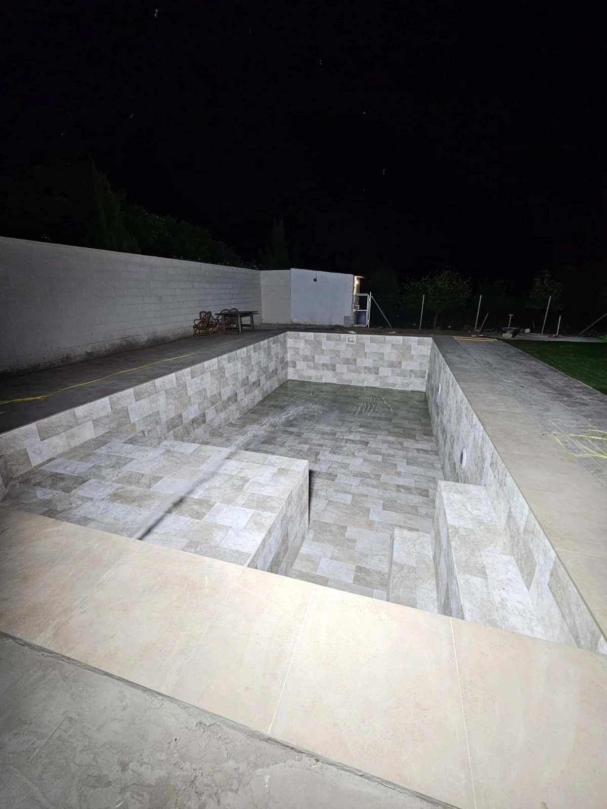 Piscina rectangular de hormigón vacía, de noche, junto a una carretera asfaltada y un muro