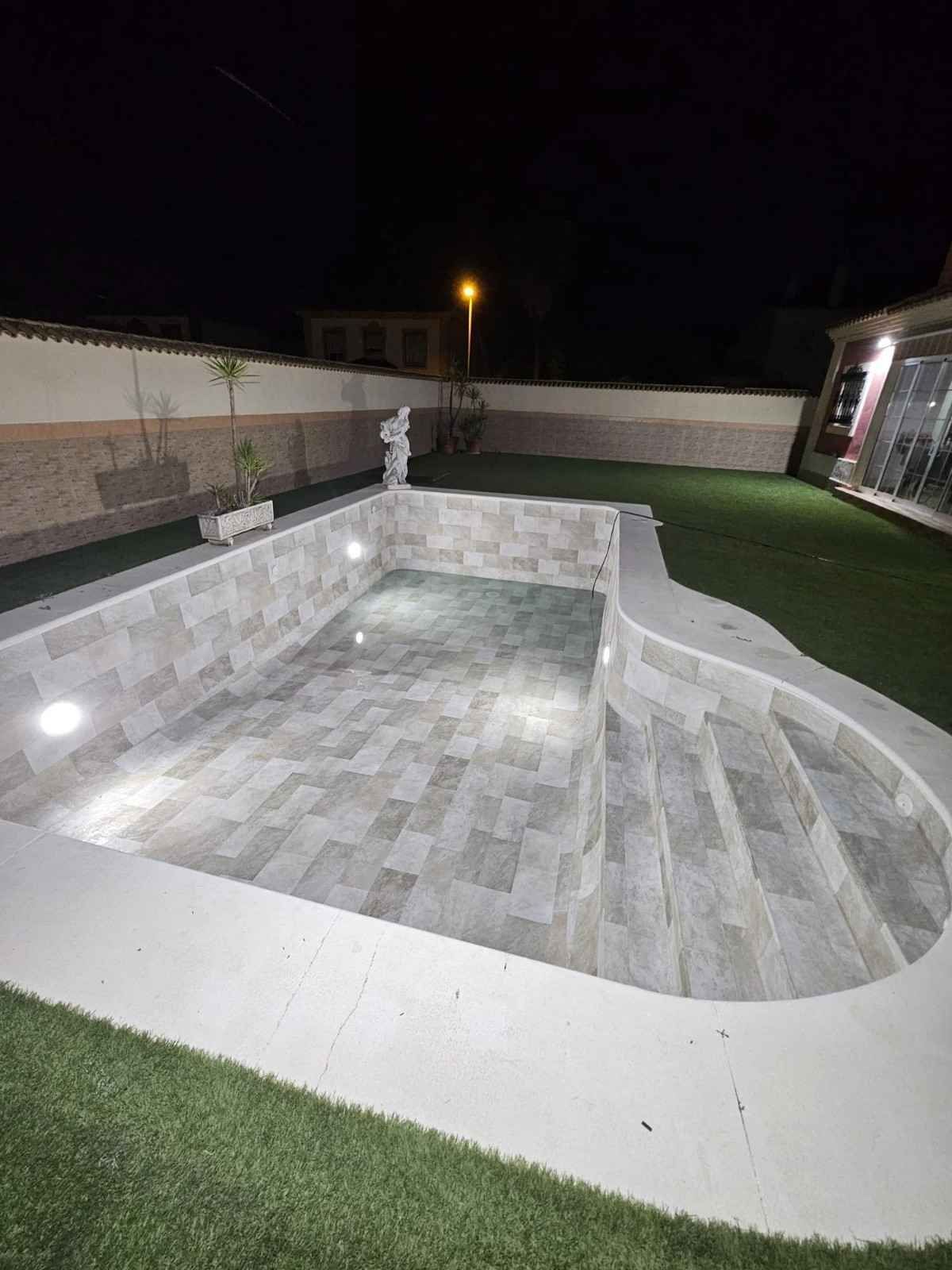 Piscina enterrada de azulejos vacía o spa de noche, junto a un césped y luces de patio.