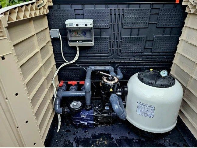 Zona de equipos de piscina al aire libre con bomba, caja de control y depósito de filtro blanco sobre una alfombrilla negra