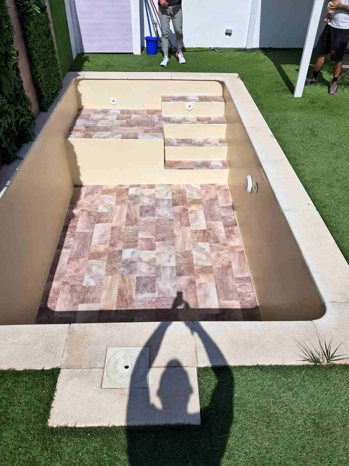 Piscina vacía enterrada en un patio trasero, con paredes color arena y suelo de azulejos, vista desde arriba.