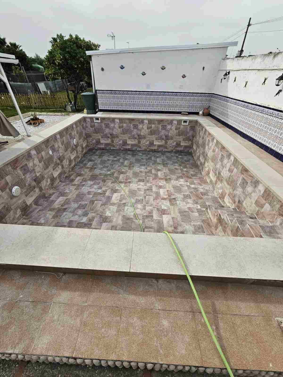 Piscina vacía en una azotea embaldosada o patio interior, con muros bajos alrededor y una manguera en primer plano