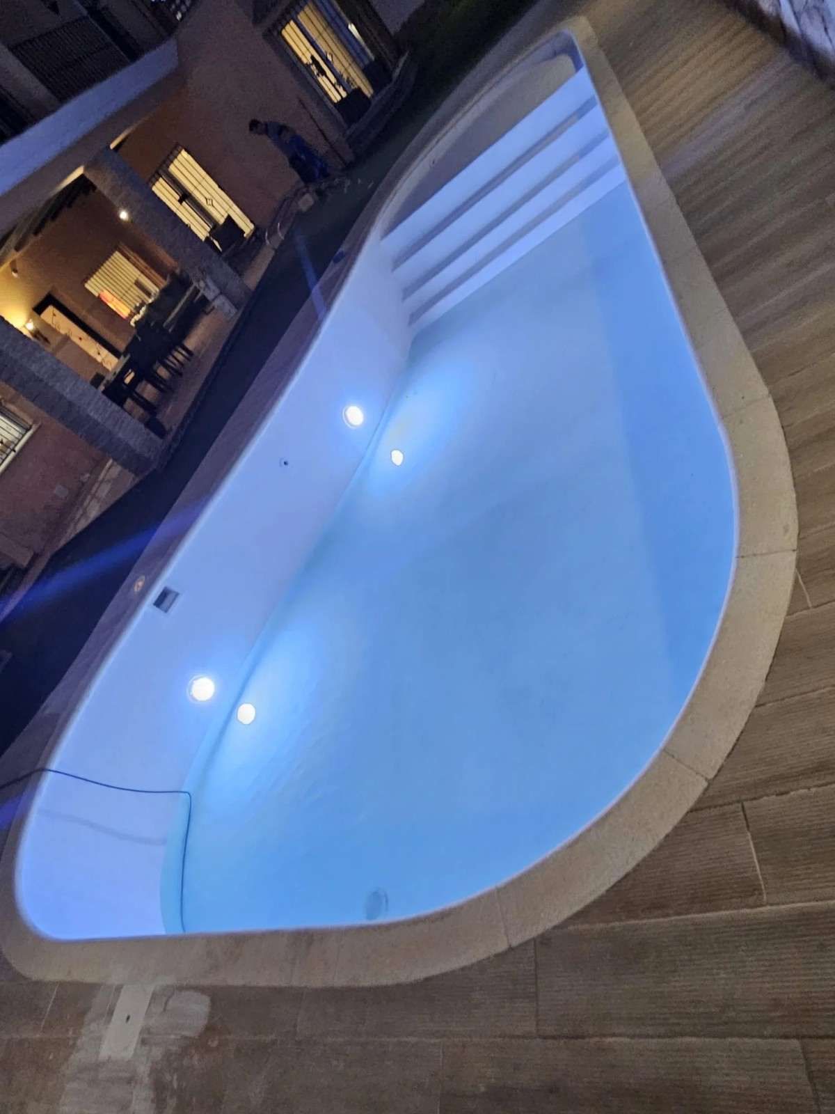 Piscina exterior iluminada en azul por la noche, junto a una terraza de madera y las ventanas iluminadas de un edificio