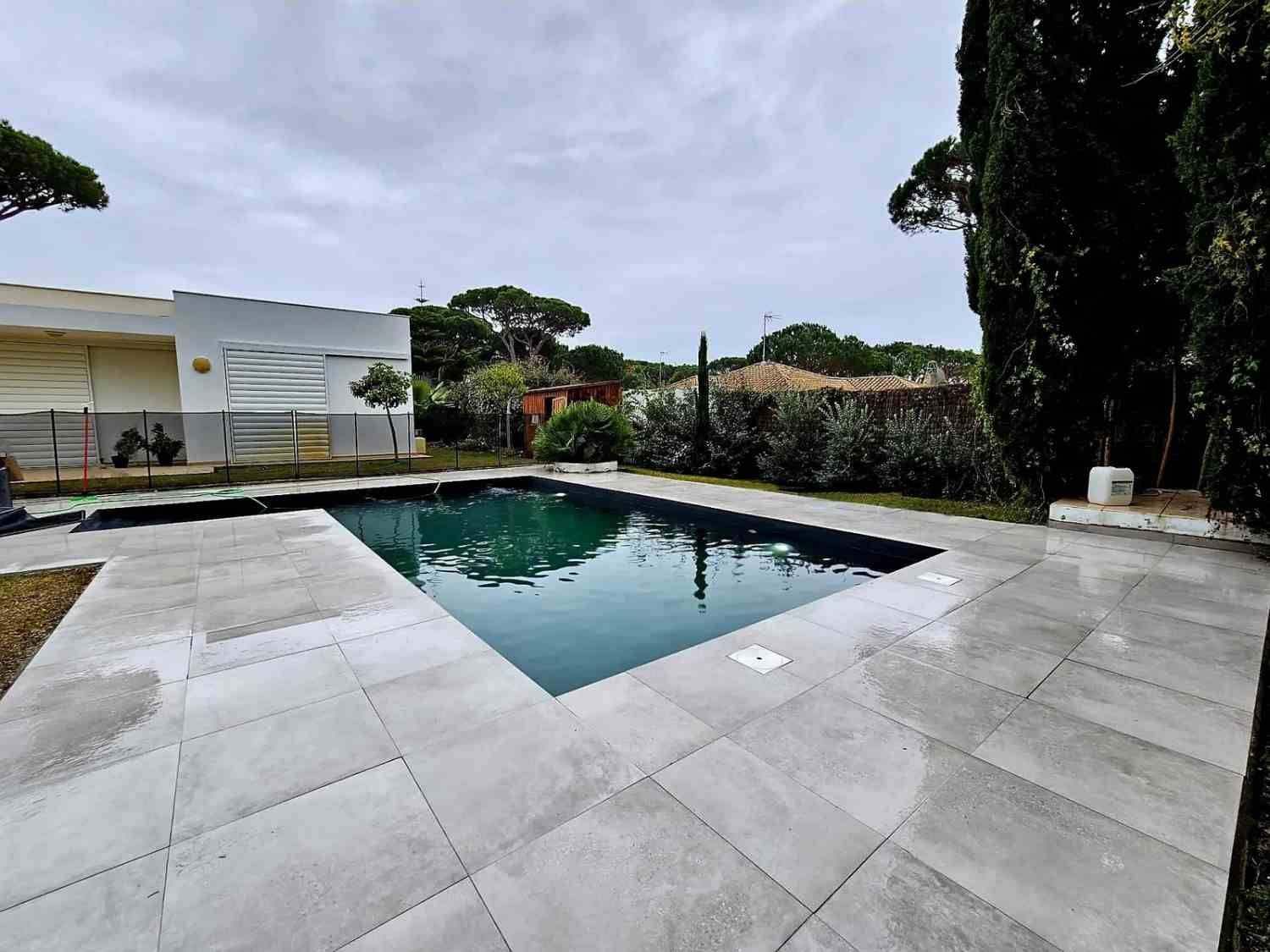 Jardín trasero moderno con una piscina rectangular, un amplio patio pavimentado y vegetación alrededor bajo un cielo nublado