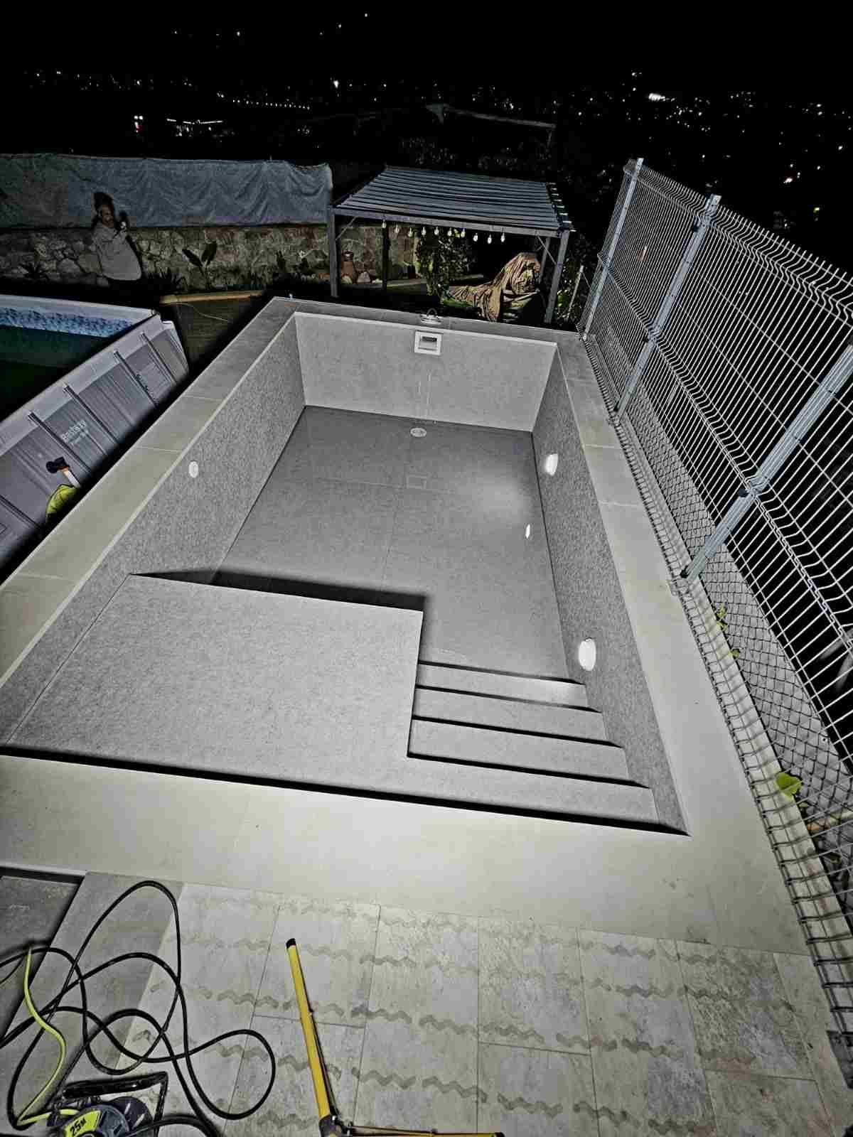 Piscina rectangular de hormigón vacía por la noche, vista desde arriba, con una valla y unas escaleras a la derecha.