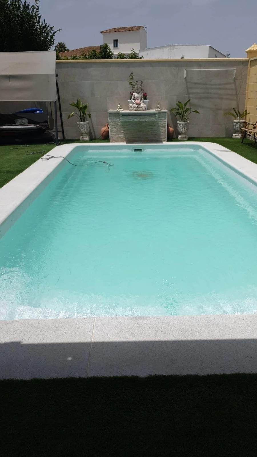 Piscina turquesa en un patio trasero, con un muro de piedra, plantas en macetas y una fuente decorativa al fondo