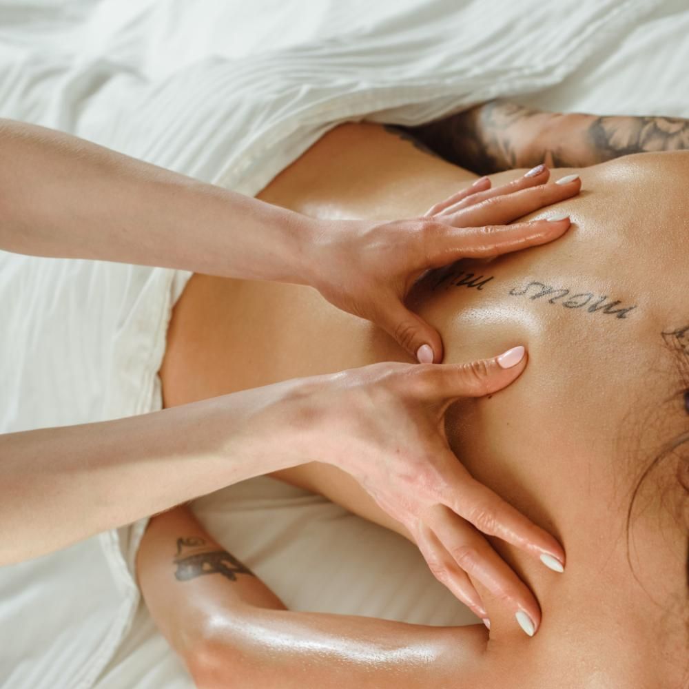 Manos masajeando la espalda de una persona, tatuajes visibles. Sábanas blancas.