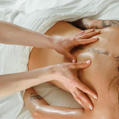 Manos masajeando la espalda de una persona, tatuajes visibles. Sábanas blancas.