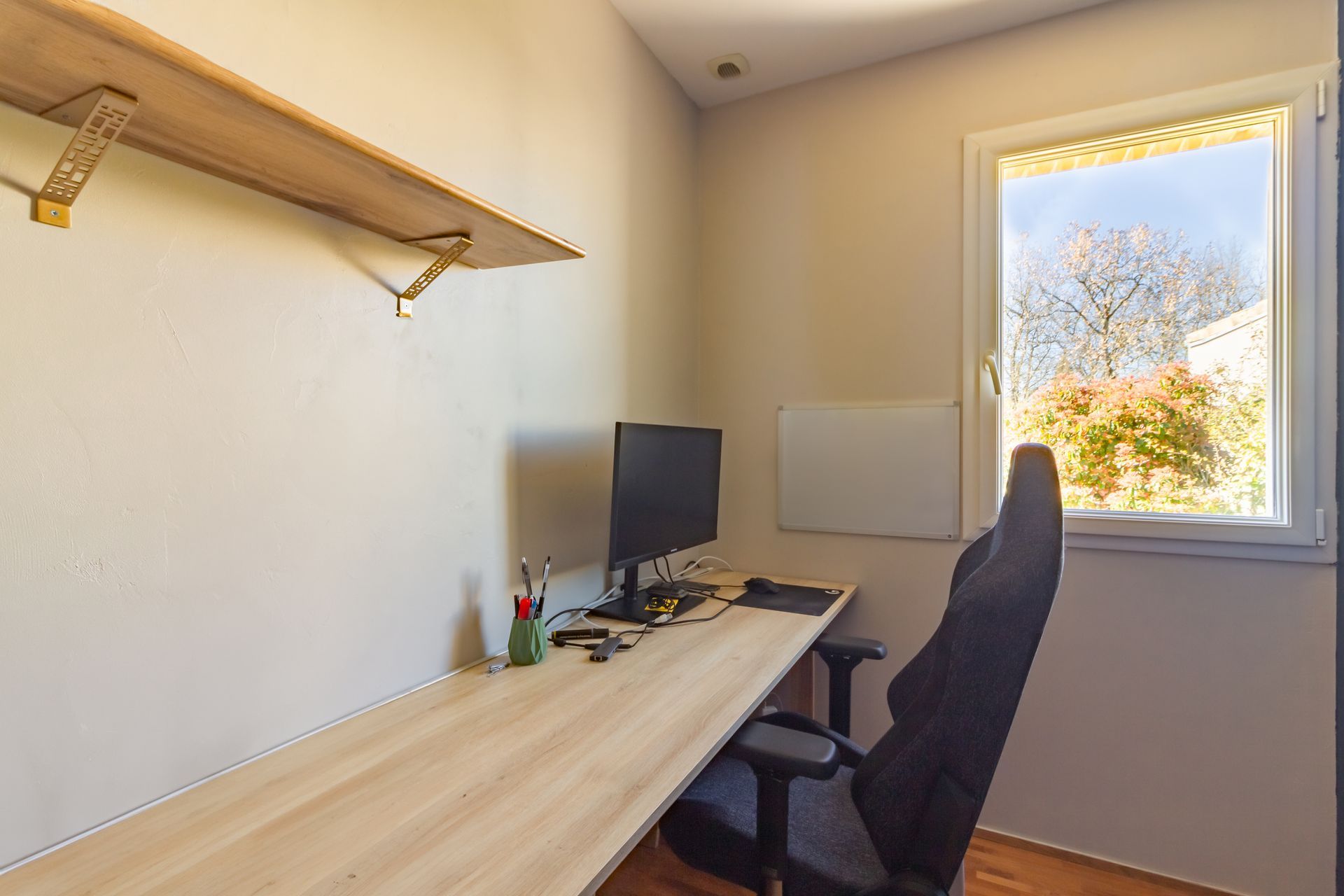 Bureau à domicile avec bureau, ordinateur et chaise près d'une fenêtre. Une étagère est placée au-dessus du bureau.